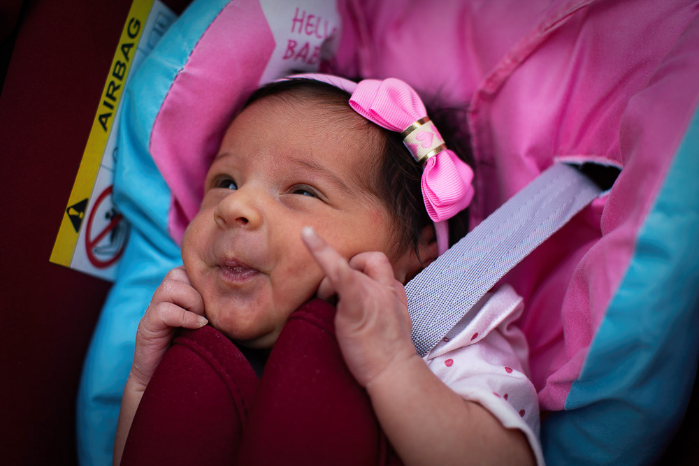Anya in a pink car seat with a bow on her head, smiling. Águas Santas, Portugal. 2025 © Pedro Rodrigues